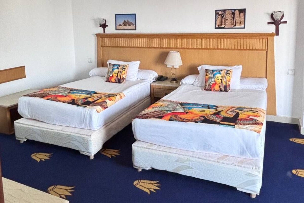 A hotel room with two single beds, a wooden headboard, a nightstand with a lamp, and a blue carpet with leaf patterns.