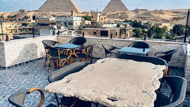 A rooftop terrace with a view of the pyramids, wicker chairs, a table with a white tablecloth, and patterned floor tiles. - A pyramids view (Giza)