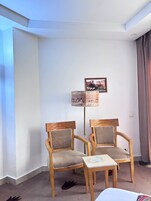 A room with two wooden chairs and a small table, a picture on the wall, and a window with curtains.