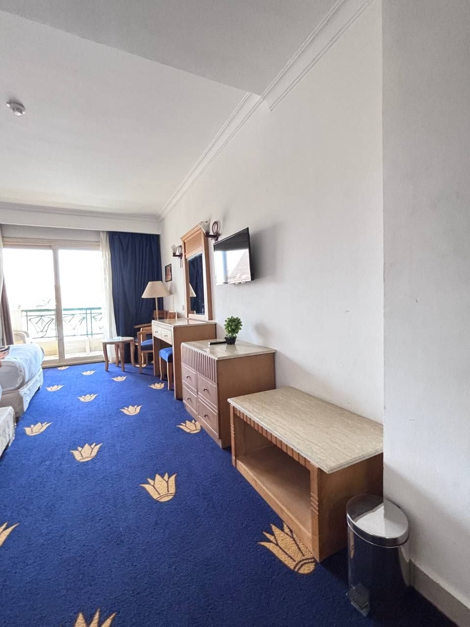 A hotel room with a bed, a desk, a chair, a small table, a trash bin, and a TV on the wall.