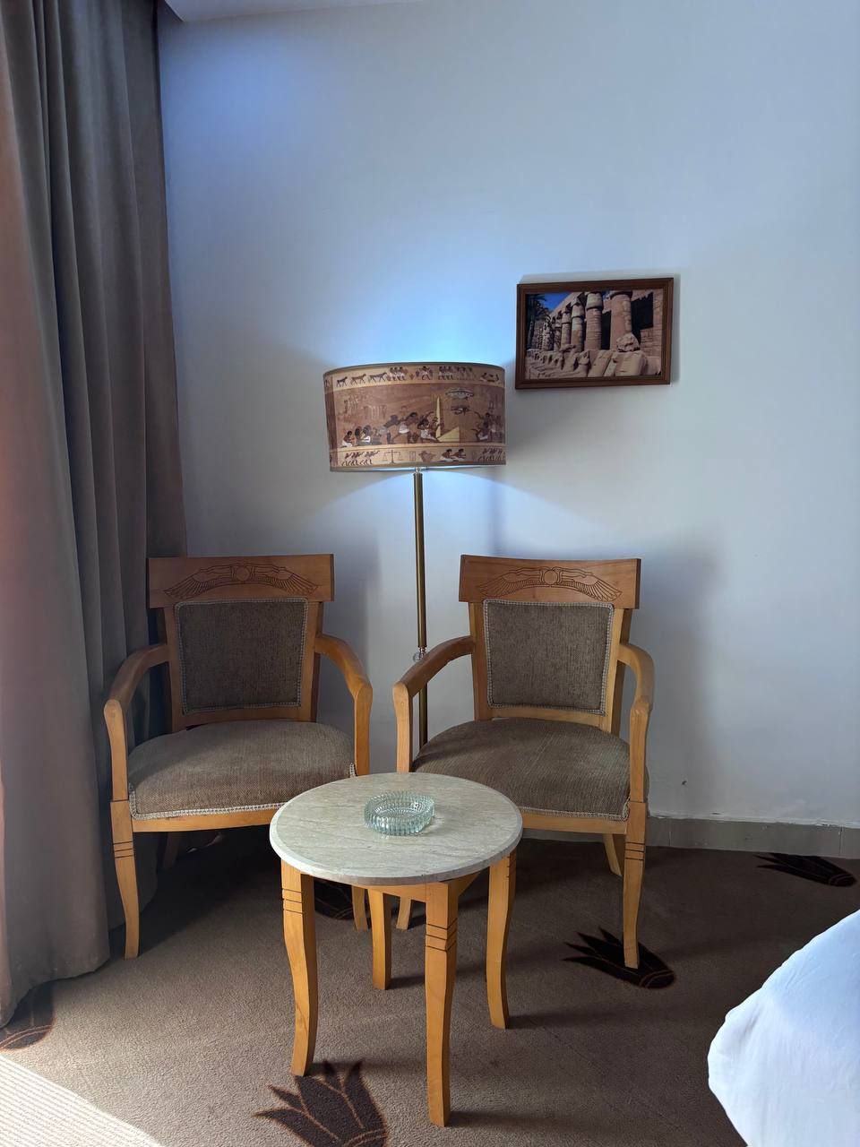 A room with two wooden chairs and a small round table, a lamp with a painted shade, and a framed picture on the wall.