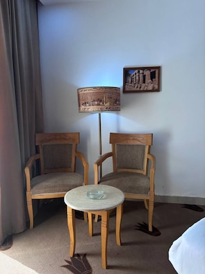 Et værelse med to træstole og et lille rundt bord, en lampe med en malet skærm, og et billede i ramme på væggen.