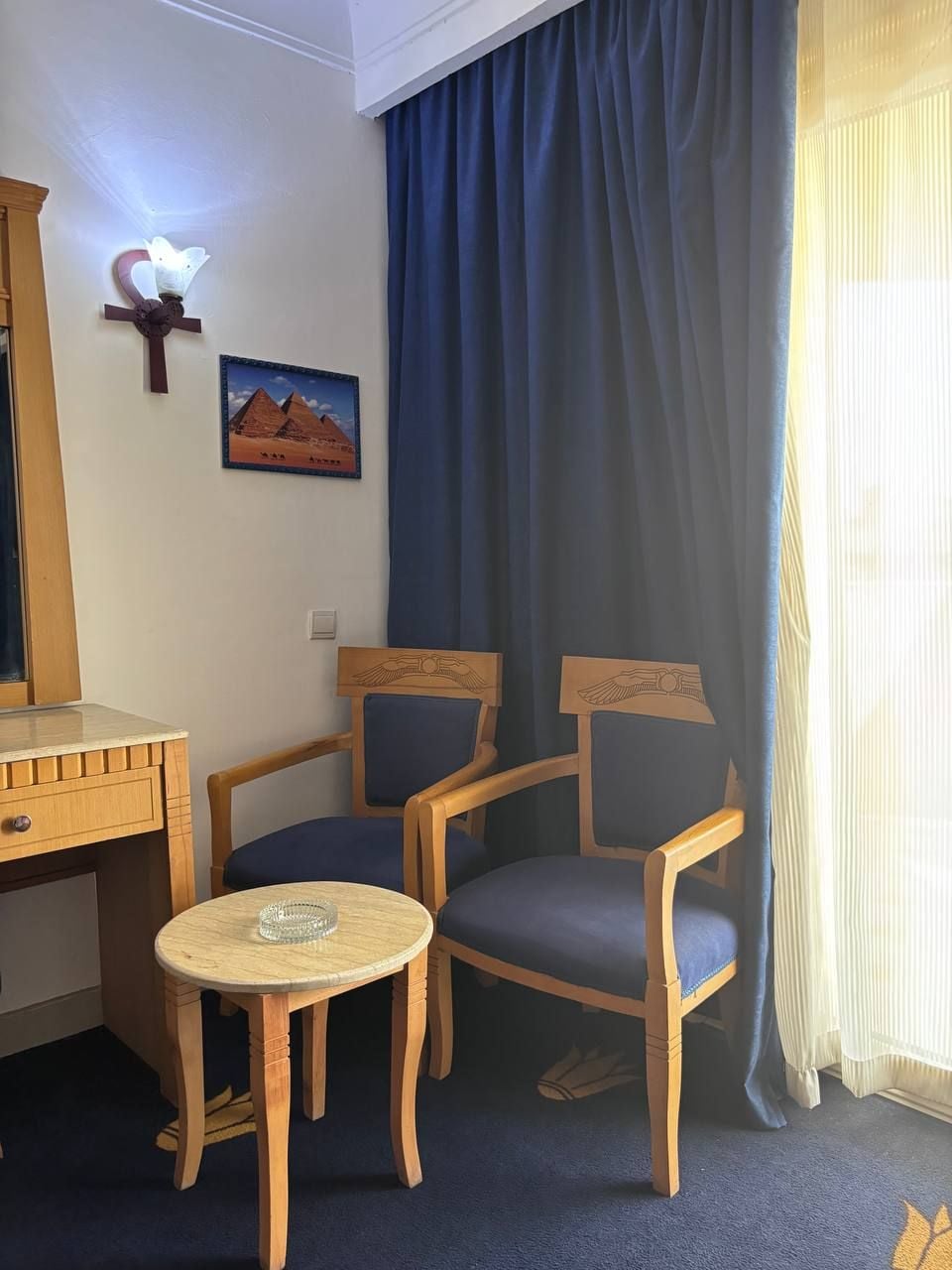 A room with a desk, two chairs, a small round table, a wall-mounted lamp, a framed picture of pyramids, and a window with curtains.