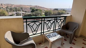 Un balcón con dos sillas, una mesita pequeña, suelo de baldosas y vistas a colinas y edificios distantes.