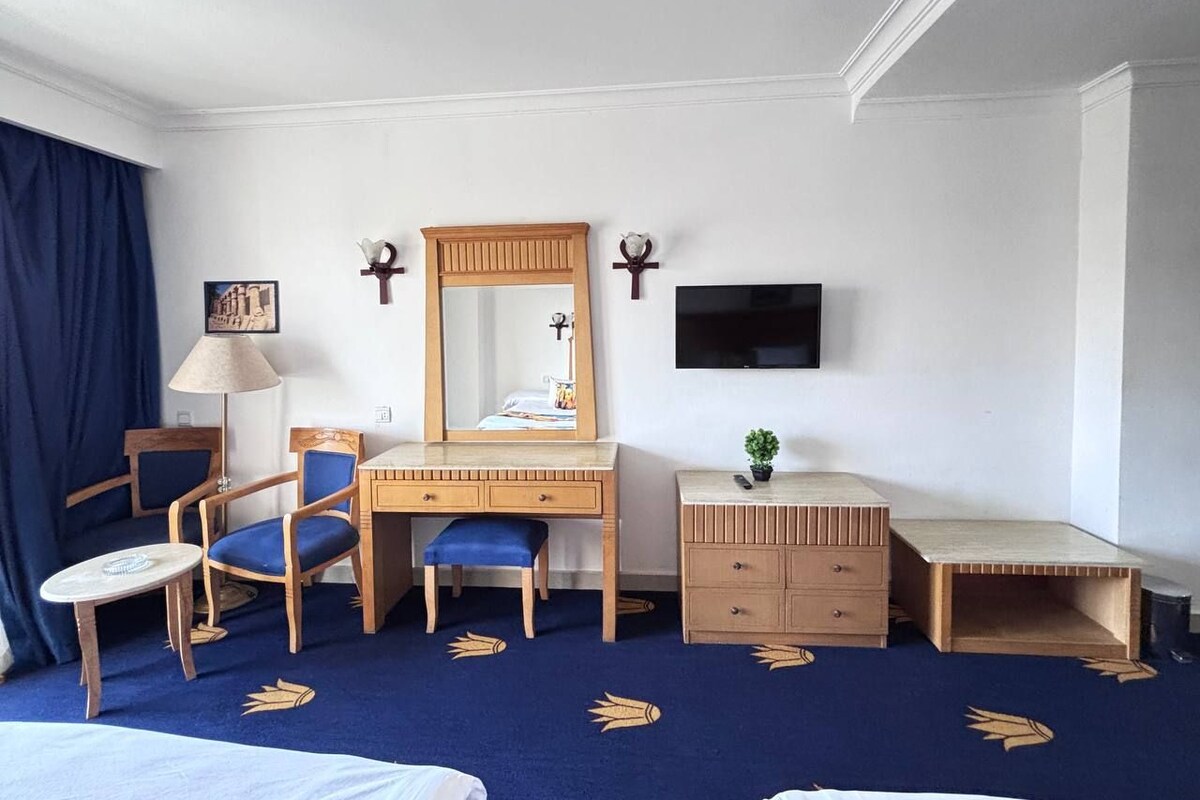 A hotel room with a bed, a desk with a chair, a dresser, a TV mounted on the wall, and a small plant on the dresser.