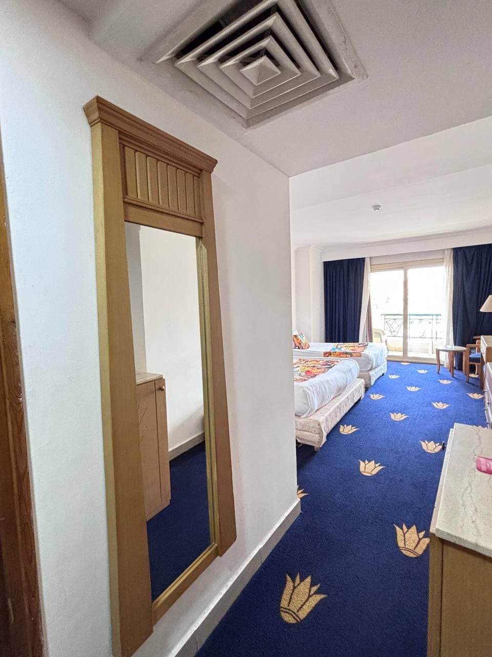 A hotel room with a large mirror, a bed with a floral carpet, a desk with a lamp, and a view of the outside through a window.