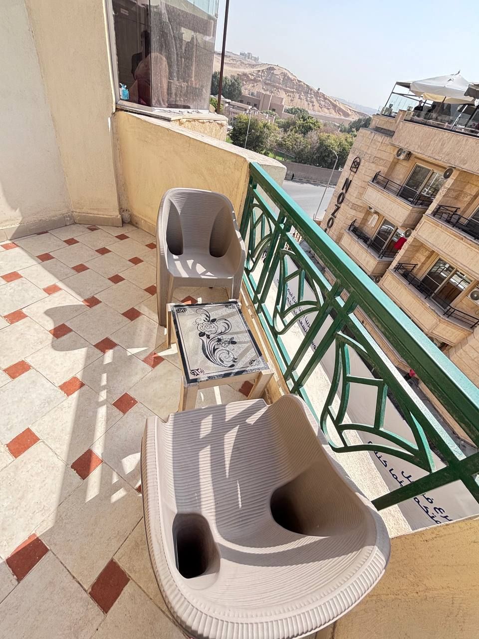 Un balcon doté d’un sol carrelé, d’une chaise en plastique, d’une petite table ornée d’un carreau décoratif et d’une rambarde verte.