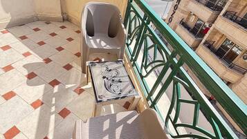 Un balcón con suelo de baldosas, una silla de plástico, una mesita con un azulejo decorativo y baranda verde.