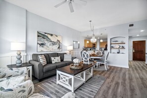 A modern living room with a gray sofa, a coffee table, and a dining area with a chandelier. - Origin 1211 - One Bedroom - Spacious Partial Gulf View (Panama City Beach)