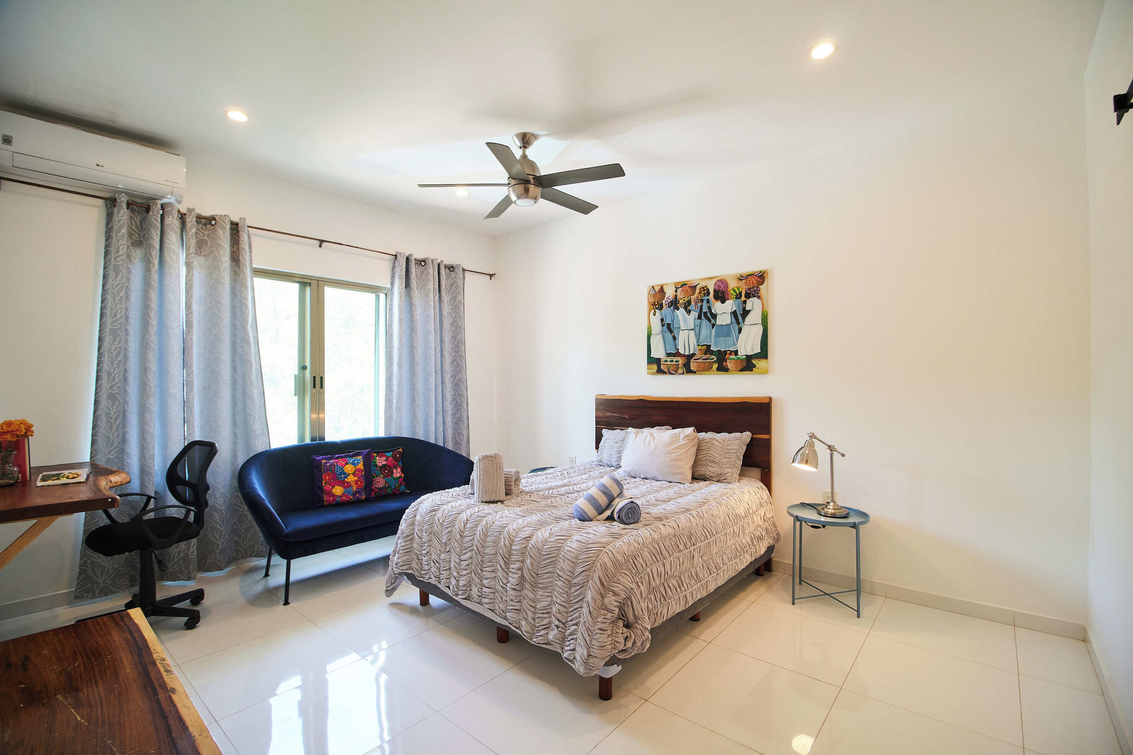 Une chambre à coucher comprenant un lit, un bureau, une chaise, un canapé, un ventilateur de plafond et un tableau accroché au mur.