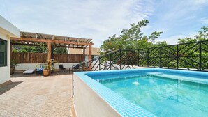 A swimming pool with a tiled edge, a wooden pergola, and a covered seating area with chairs and potted plants.