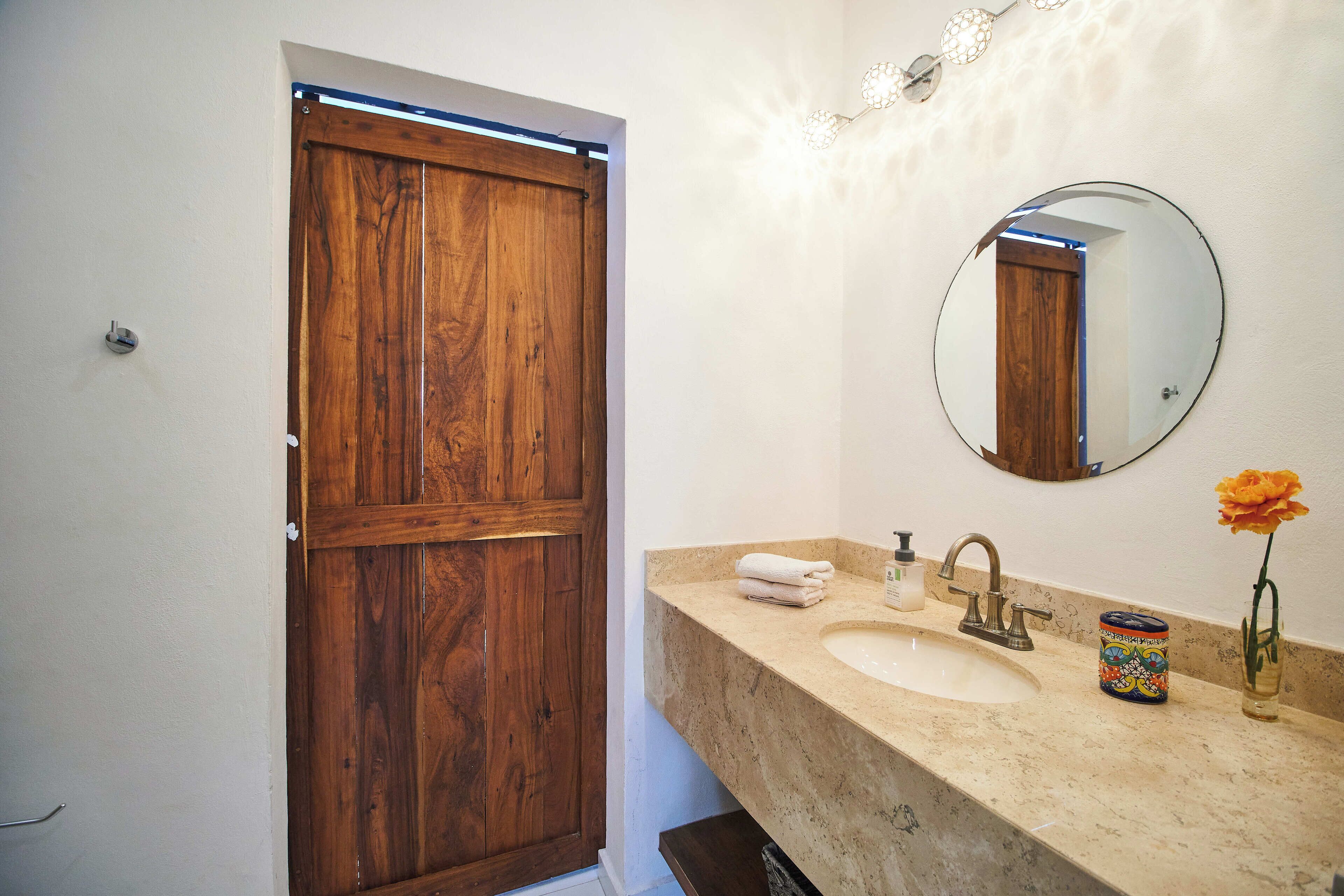 Une salle de bain avec une porte en bois, un miroir rond, un lavabo et un vase avec une fleur.