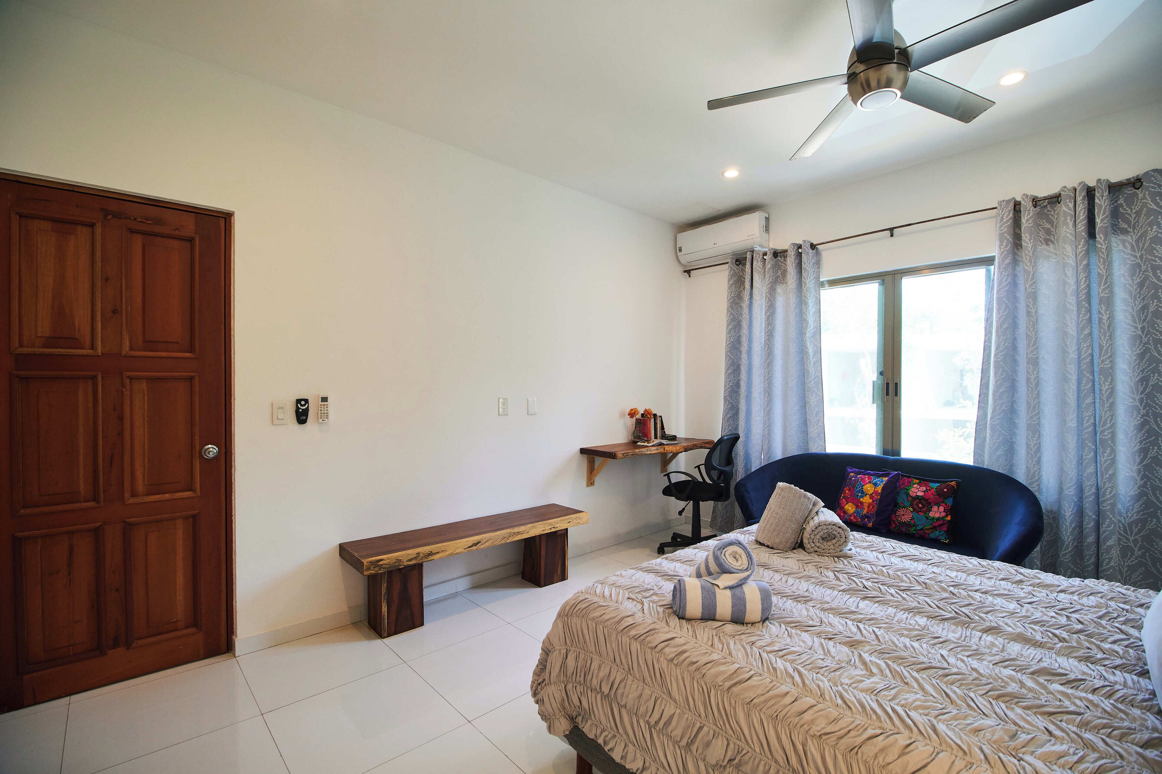 A bedroom with a bed, a desk, a chair, a bench, and a ceiling fan.