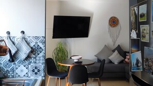 En moderne stue med et fladskærms-tv, et rundt spisebord, sorte stole, en grå sofa og en plante ophængt på væggen.