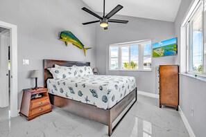 Quarto com cama, ventoinha no teto, mesa de cabeceira, cômoda, arte de parede com tema de peixes, janela com vista e pintura na parede.