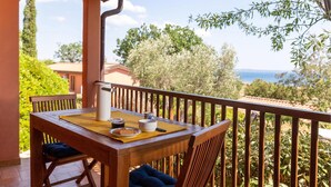 Una mesa de madera con una jarra blanca, un tazón y un plato con galletas en un balcón con vistas al mar y árboles.