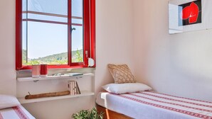 Un dormitorio con cama, una ventana con cortinas rojas, estantería con libros y una planta, y una obra de arte colgada en la pared.