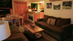 A cozy living room with a brown sofa, wooden coffee table, and a dining area with a wooden table and chairs. A kitchen is visible in the background with wooden cabinets and a countertop. - Notchtop Mountain Condo (Estes Park)
