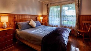 A bedroom with a wooden bed, two bedside tables with lamps, a chair, and a window with curtains. - Tarkinegrove... welcome to the wild side. (Roger River)