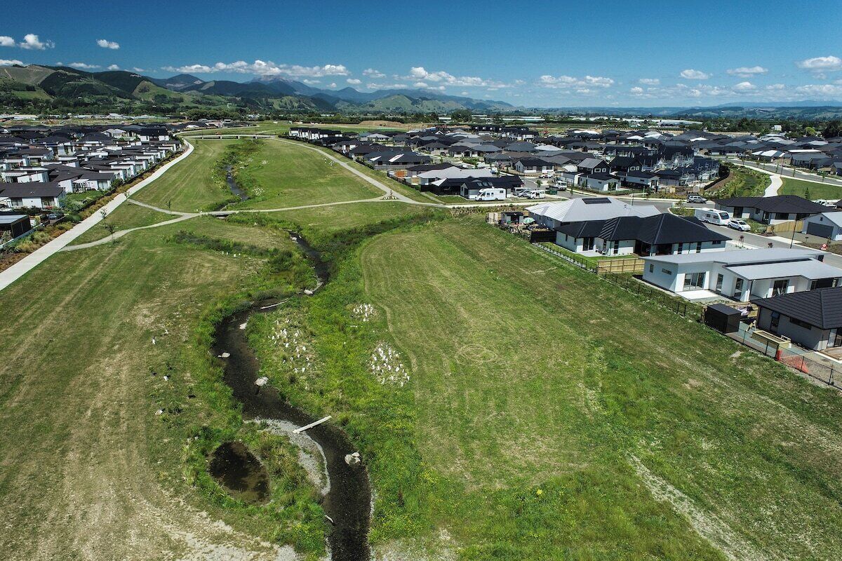 A residential area with houses arranged in rows along a curved road, surrounded by green fields and mountains in the background.