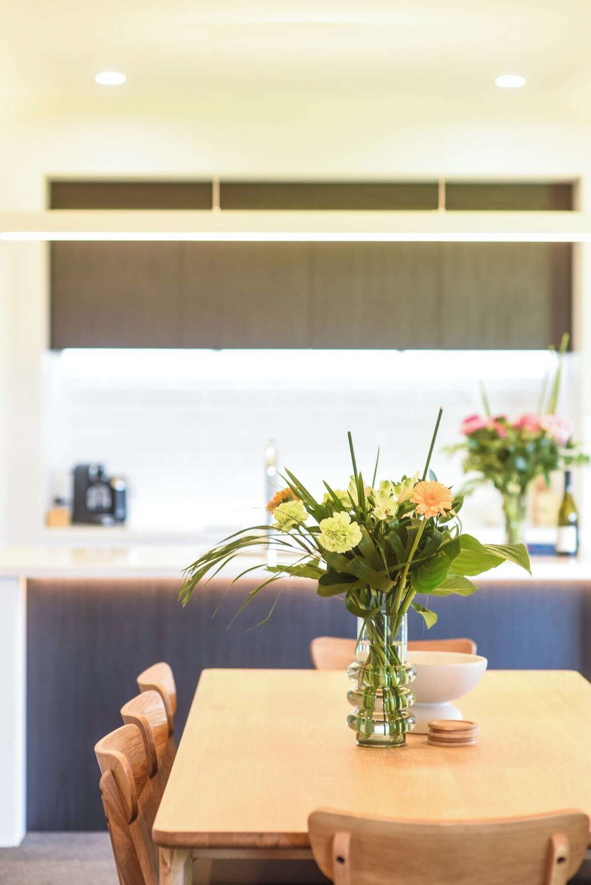 Une table à manger avec un vase de fleurs, des chaises en bois et une cuisine en arrière-plan.