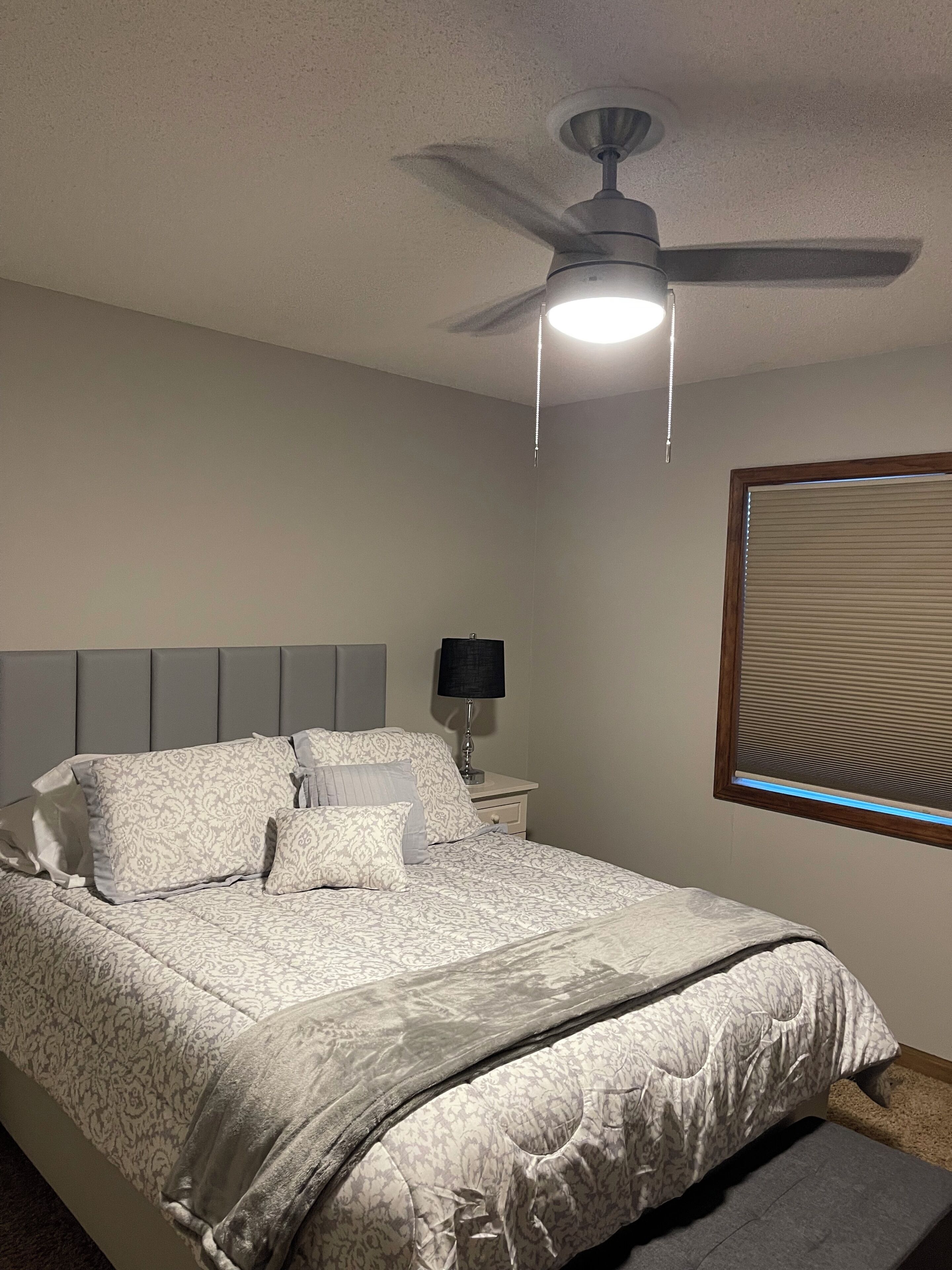 A bedroom with a bed, a nightstand, a lamp, a ceiling fan, and a window with blinds.