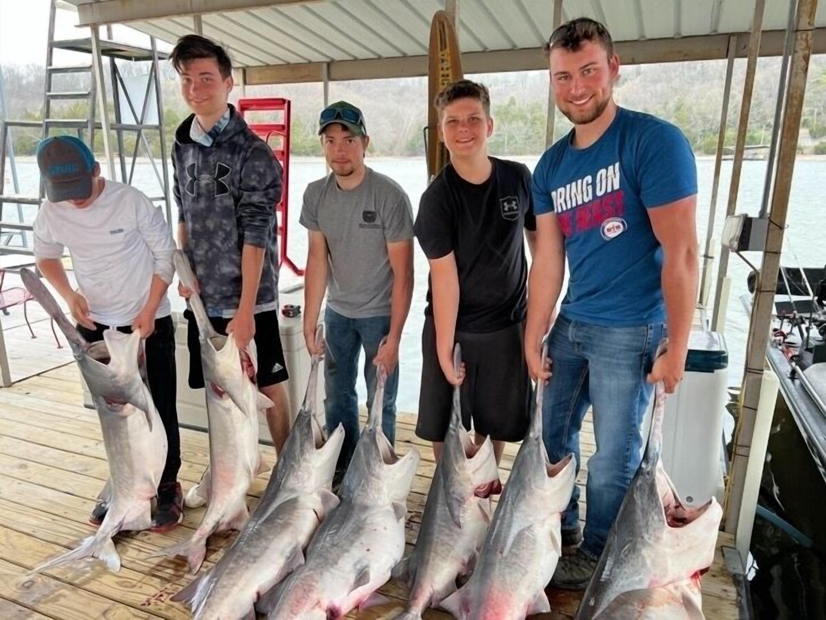 Un grupo de personas en un muelle de madera sostienen peces grandes, con un bote y equipo de pesca al fondo.