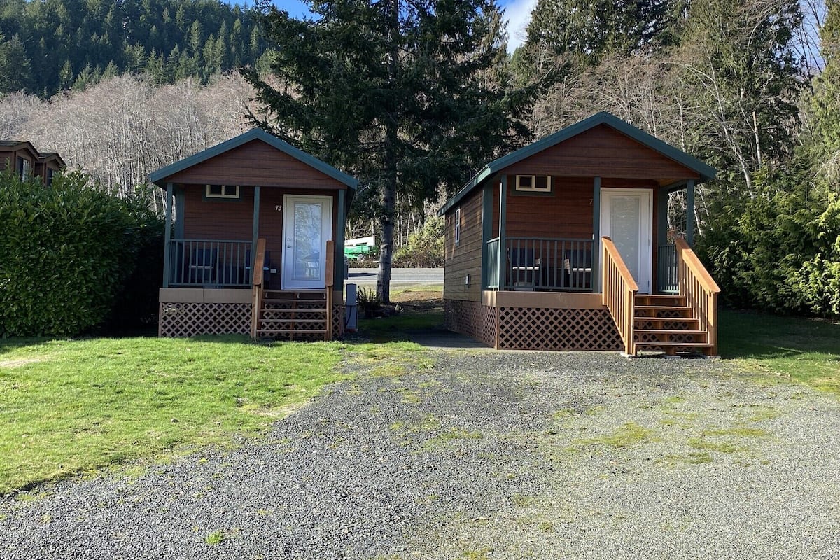 Deux cabanes en bois, dotées d’un porche et d’un escalier menant à la porte, sont entourées d’un chemin de gravier et de verdure.