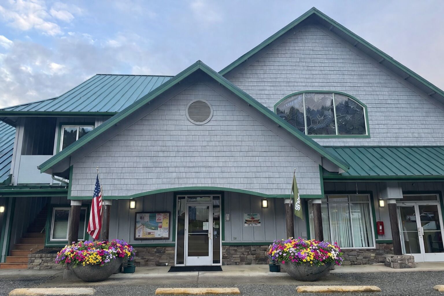 A two-story building with a green roof and a large entrance door. There are two flower planters in front of the entrance.