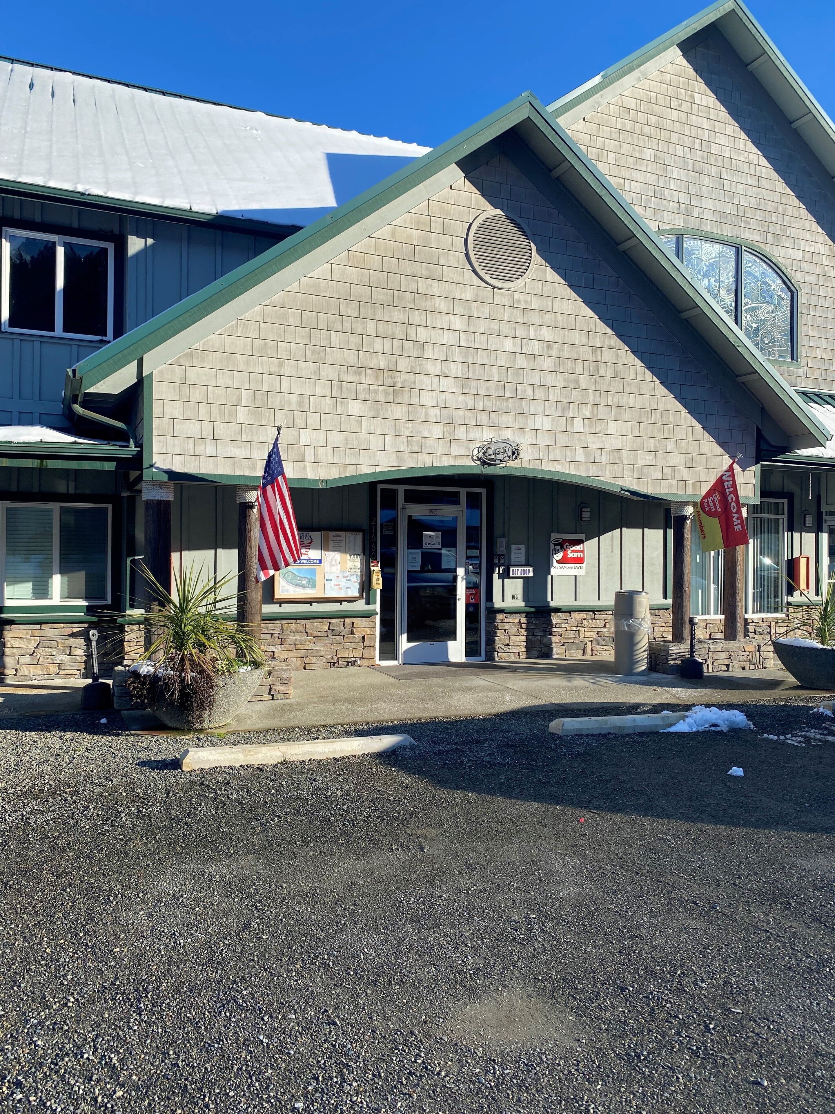 A building with a brick facade, a green roof, and an American flag. There are two flags, one red and one yellow, and a welcome sign.