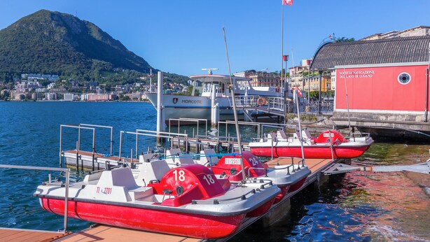 A marina with several red and white boats docked, a red building with signage, and a mountainous backdrop. - Dandy House Few Min From Lake (Lugano)