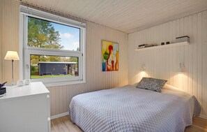 A bedroom with a large window, a bed with a patterned bedspread, a bedside table with a lamp, and a colorful abstract painting on the wall. - 3 bedroom awesome home in Glesborg (Glesborg)