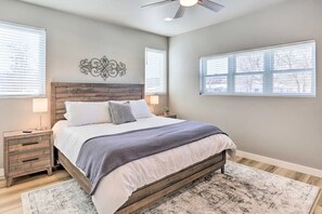 Un dormitorio con cabecera de madera, cama con ropa de cama blanca y gris, mesita de noche con lámpara y un ventanal con persianas.