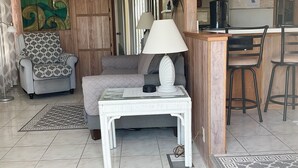 A living room with a sofa, a patterned armchair, a small table with a lamp, a kitchen area with a refrigerator, and a patterned rug. - ISLE BE BACK To The Sunny Keys (Cudjoe Key)