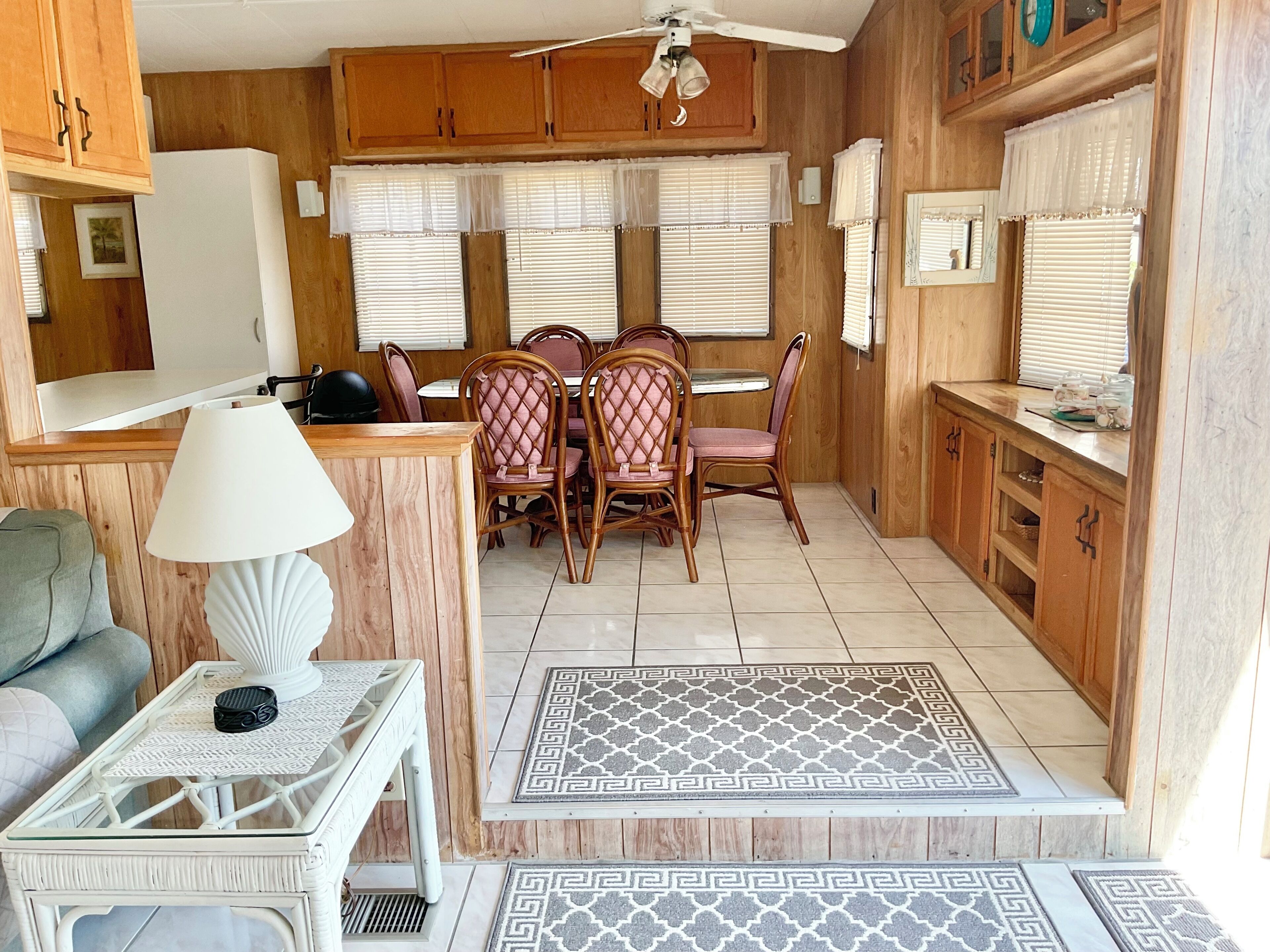 A compact kitchen with wooden cabinets, a dining table with chairs, a white lamp, and a patterned rug.