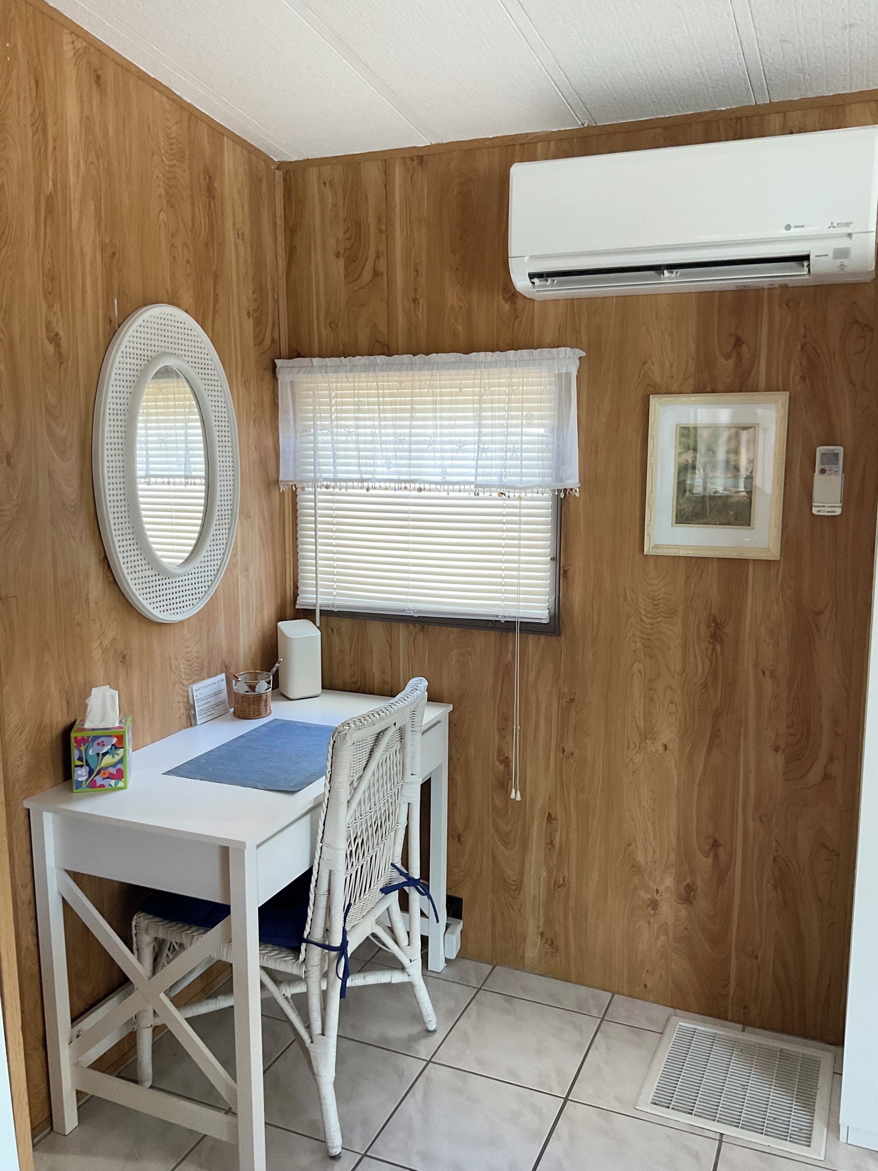 Ein Zimmer mit einer Holzwand, einer weißen Klimaanlage, einem runden Spiegel, einem Fenster mit Jalousien, einem gerahmten Bild, einem Tisch mit Stuhl und einem kleinen Korb mit Taschentuchbox.