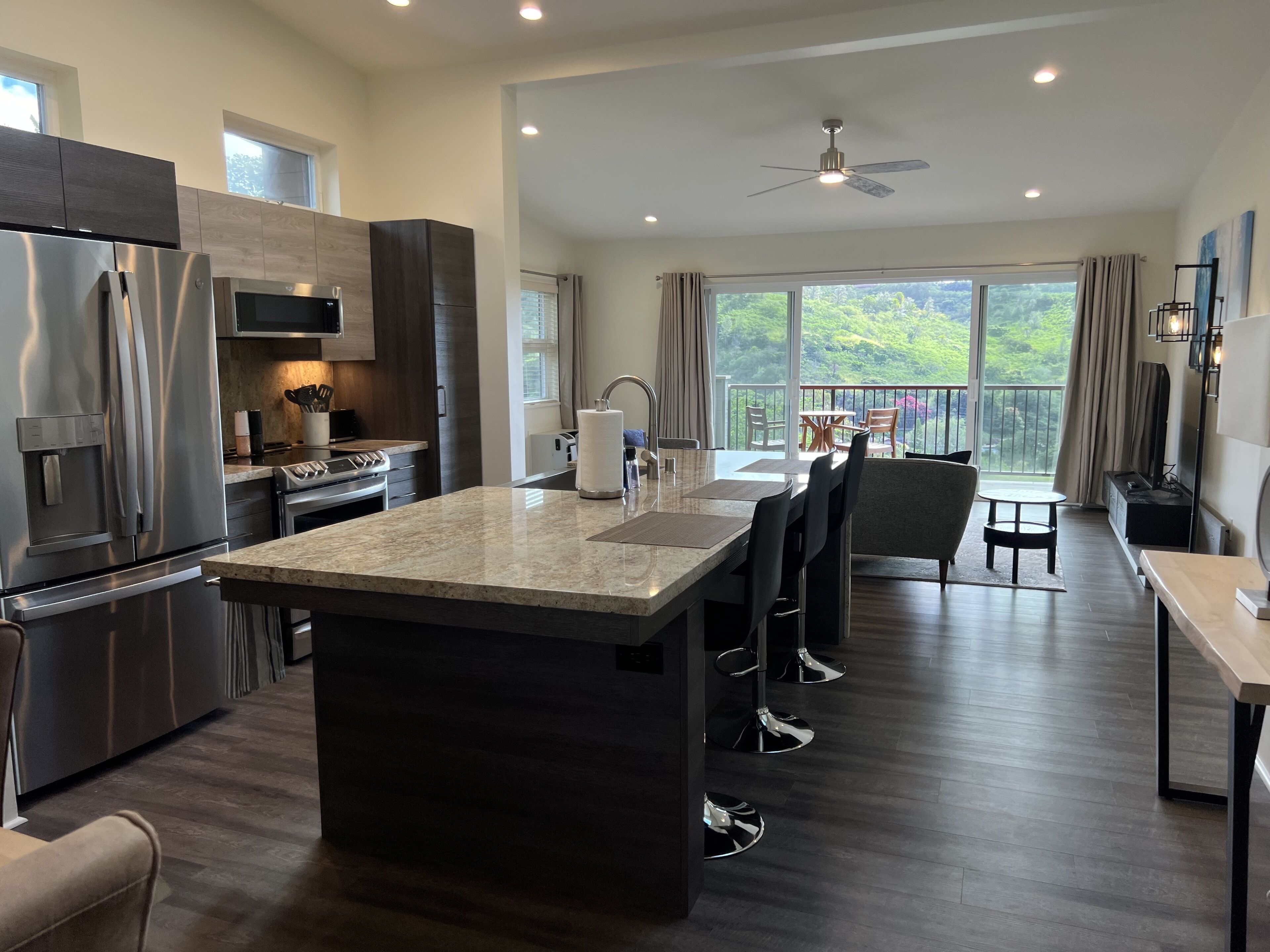 A modern kitchen with a central island, stainless steel appliances, and a dining area with a view of greenery.
