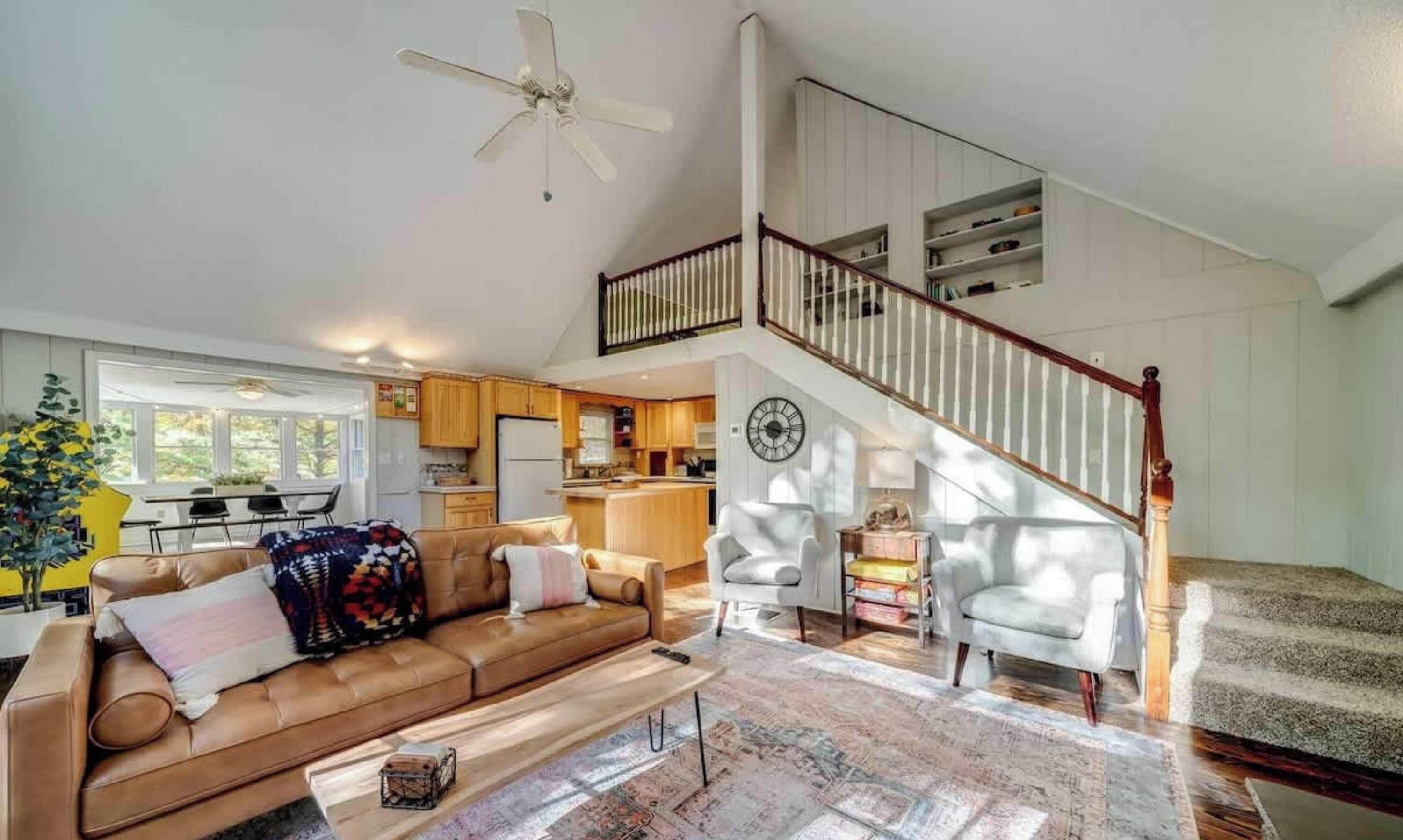 A spacious living area with a high ceiling, a staircase, and a kitchen in the background.