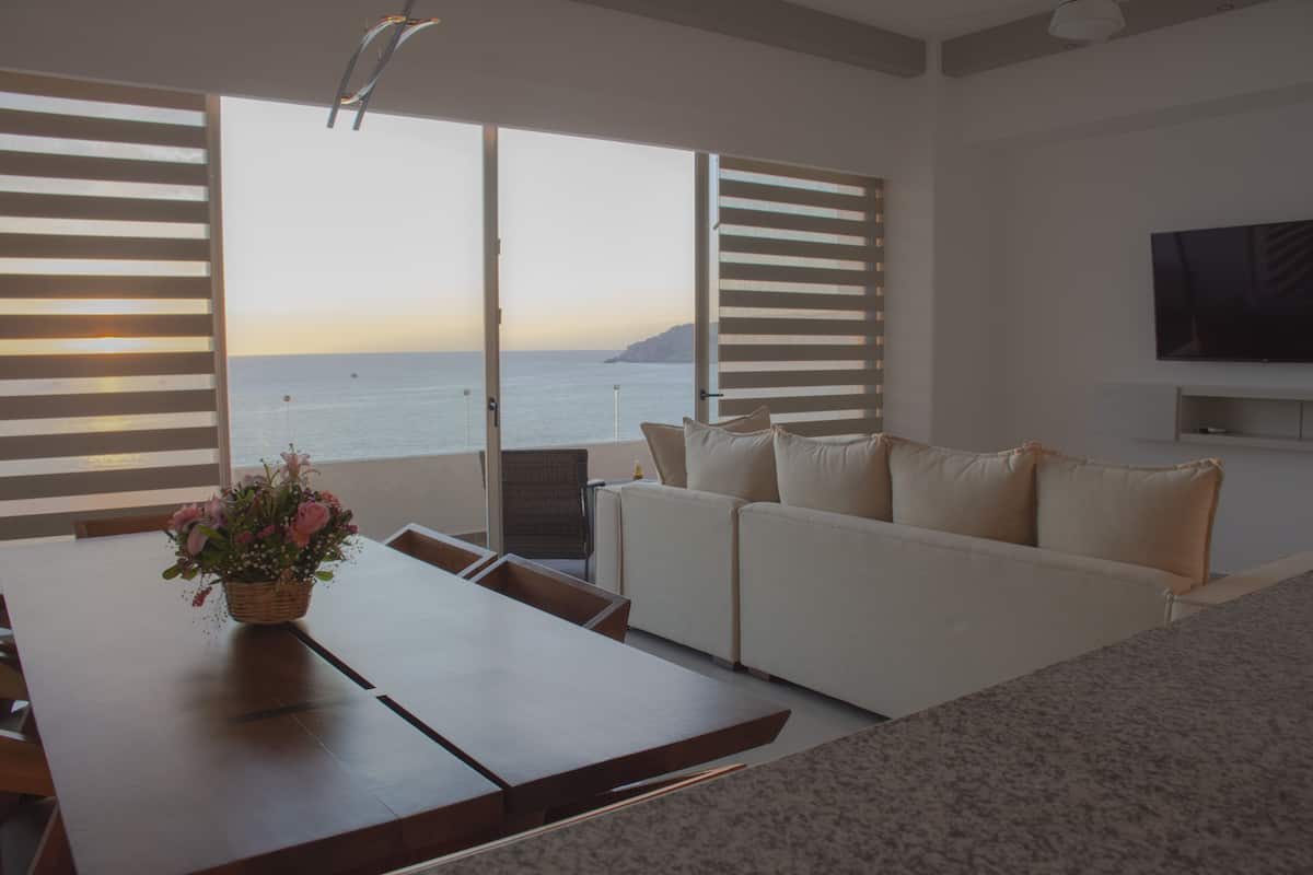 Uma sala de estar moderna com um sofá, uma mesa de jantar com um vaso de flores e vista para o mar através de grandes janelas.