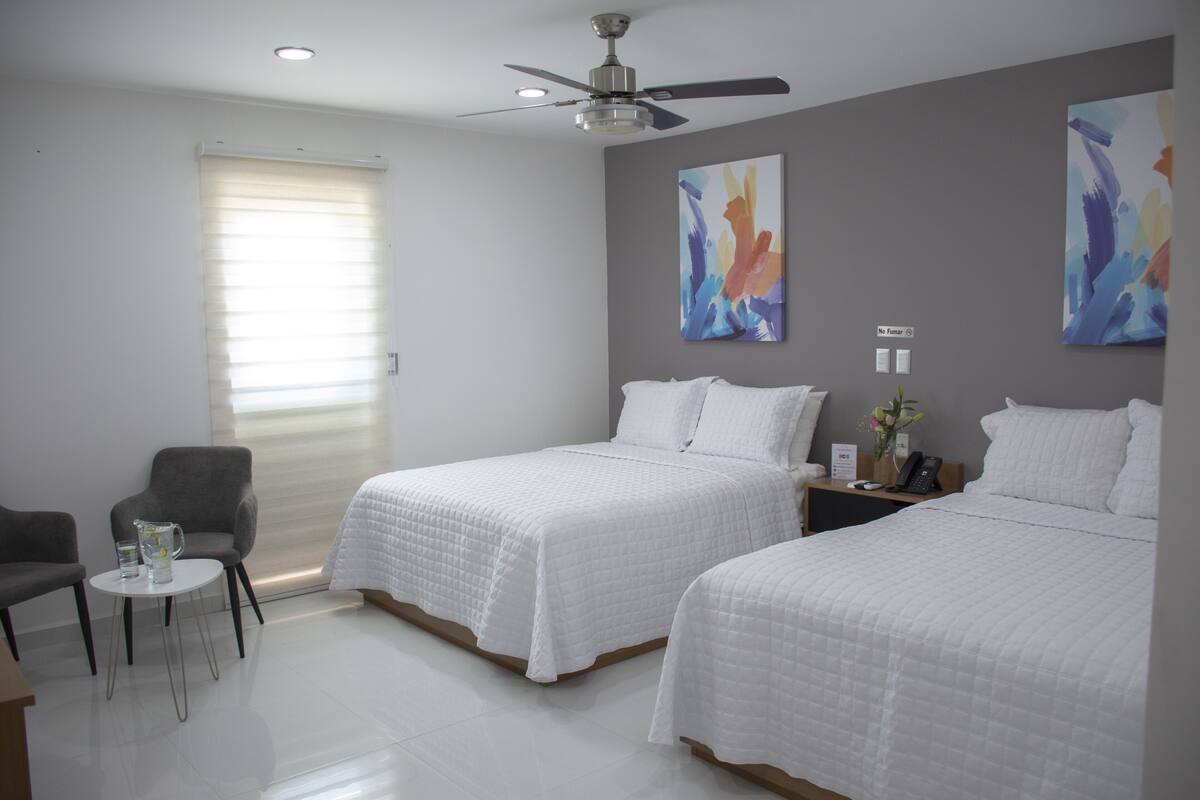 Quarto de hotel com duas camas, uma mesinha com jarra e copos, uma cadeira e quadros abstratos nas paredes.