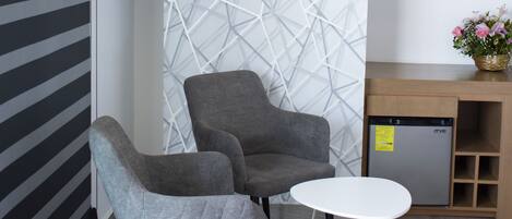 Una habitación moderna con una televisión de pantalla plana en la pared, un sillón gris, una mesita blanca pequeña y un escritorio de madera con un florero.