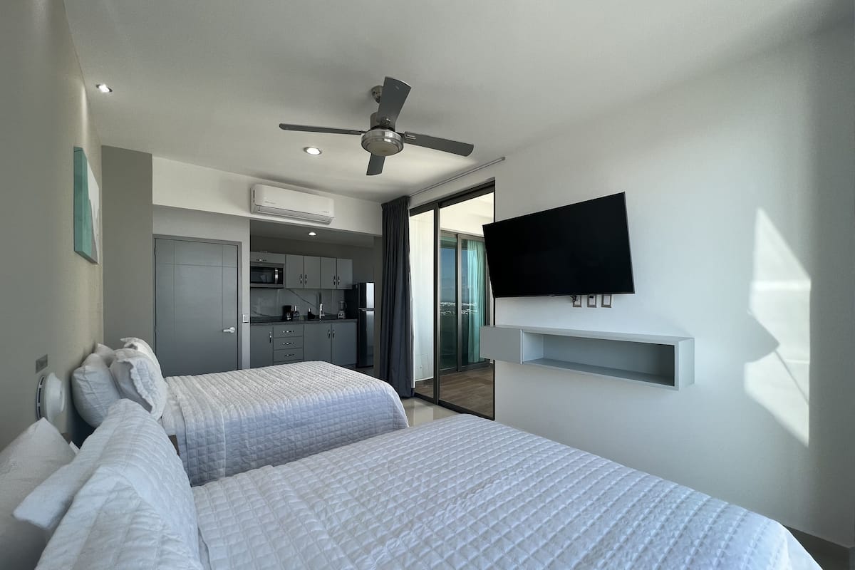 Quarto de hotel com duas camas, um ventilador de teto, televisão de tela plana, cozinha compacta e porta de vidro corria para a varanda.