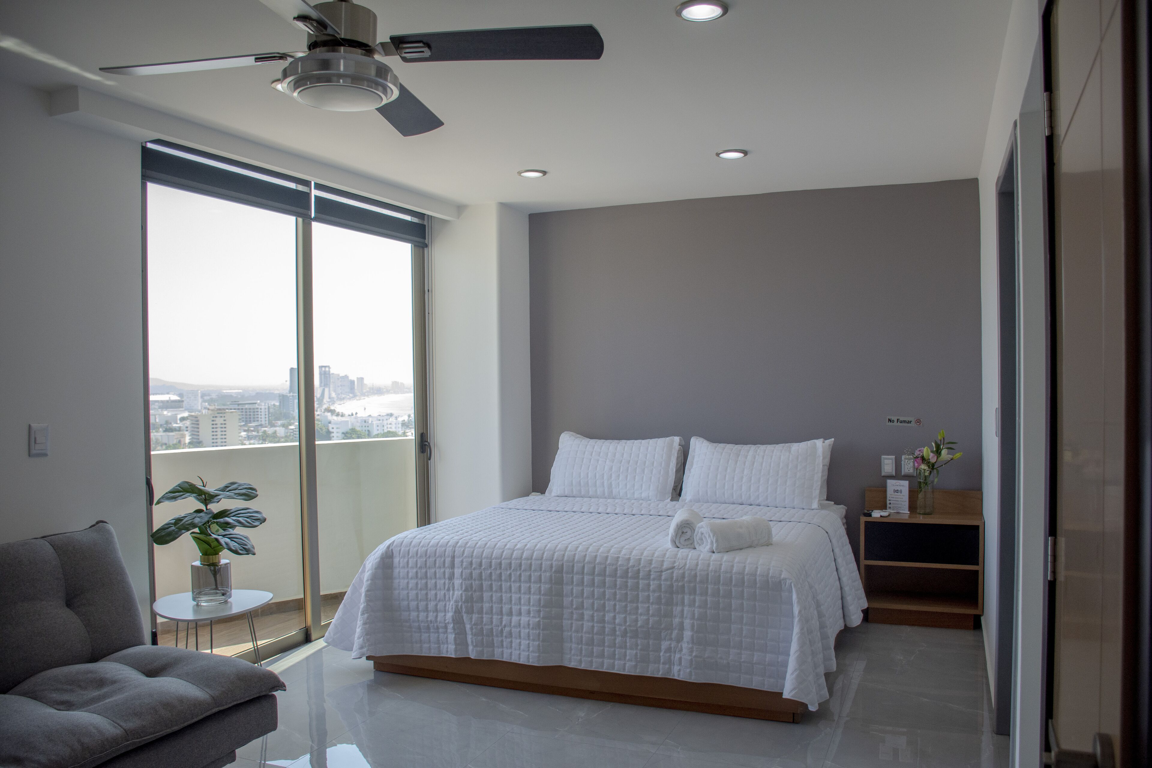 Un dormitorio moderno con una cama grande, un ventilador de techo, una mesita con una planta y vistas de la ciudad a través de amplias ventanas.