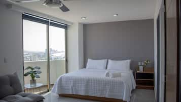 Un dormitorio moderno con una cama grande, un ventilador de techo, una mesita con una planta y vistas de la ciudad a través de amplias ventanas.