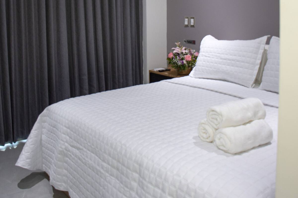 Cama bem arrumada com roupa de cama branca e duas toalhas enroladas, um ventilador de teto, uma mesa de cabeceira com um vaso de flores e uma janela grande com cortinas escuras.