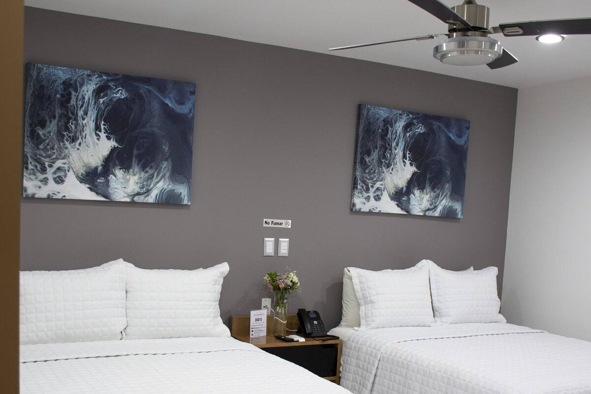 Quarto de hotel com duas camas, um ventilador de teto, obras de arte nas paredes e uma mesa de cabeceira com telefone e um vaso.