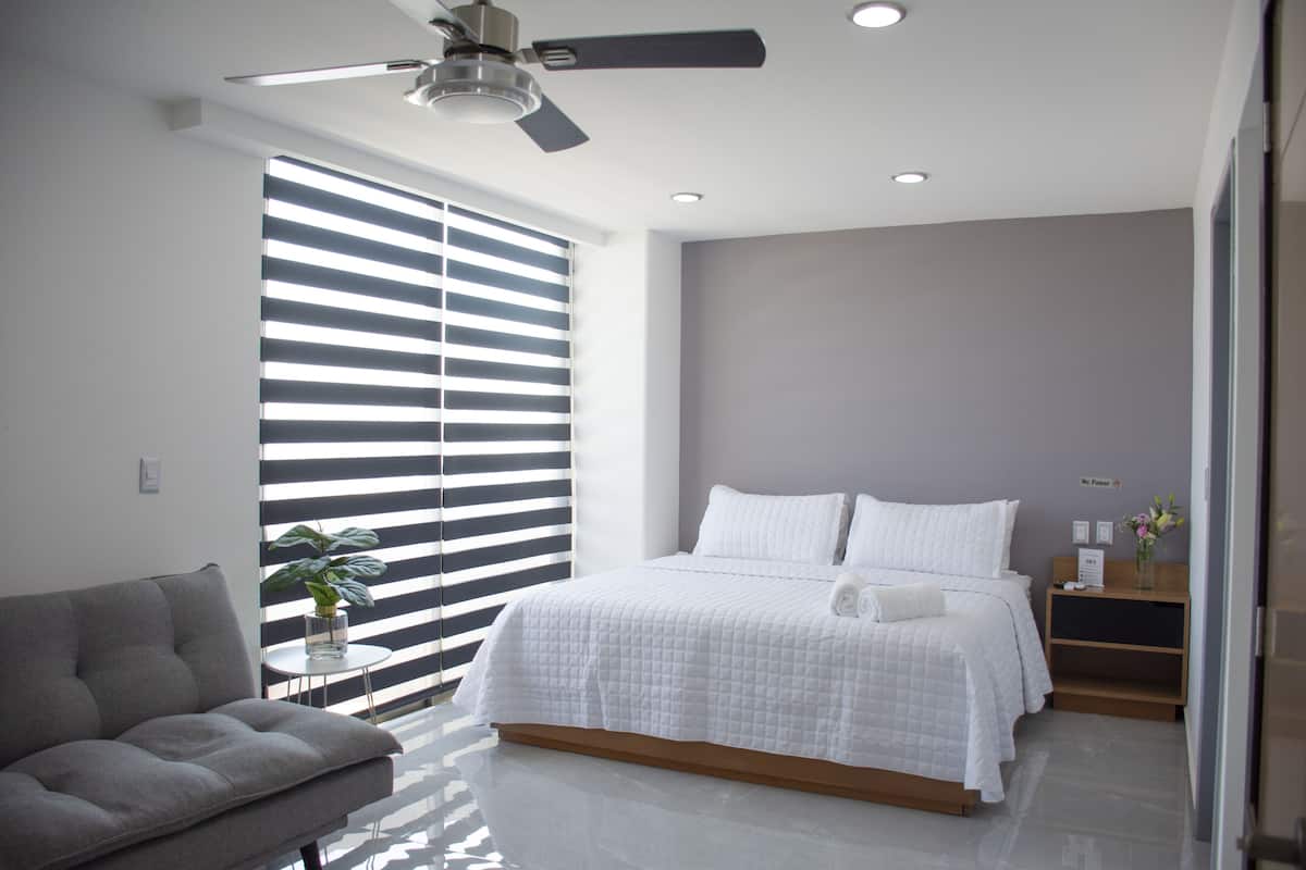 Quarto moderno com cama, sofá cinzento, uma mesinha com uma planta e um ventilador de teto.