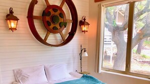 Un dormitorio con una cama, un adorno de rueda de madera grande en la pared, dos lámparas fijadas a la pared y una ventana con vistas a árboles.