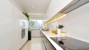A modern kitchen with a black induction cooktop, white cabinetry, and a window providing a view of the outdoors. - Prestige Sunset Carcavelos Beach - Checkinhome (Carcavelos)