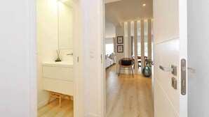 A hallway with a white door leading to a room with a wooden floor and a wicker chair. - Prestige Sunset Carcavelos Beach - Checkinhome (Carcavelos)